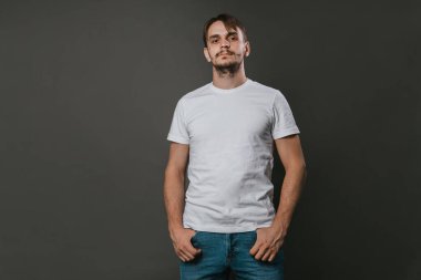 Beyaz tişörtlü ve kot pantolonlu yakışıklı bir adam gri arka planda duruyor. Stüdyo fotoğrafı. Model.