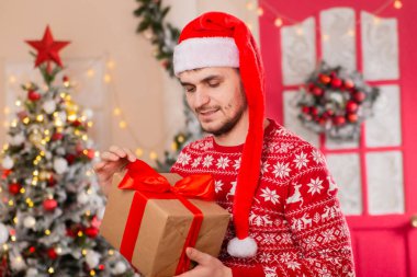 Uzun kırmızı Noel Baba şapkalı, İskandinav desenli bir süveter giyen genç bir adam bir Noel ağacının arka planına yaslanmış..