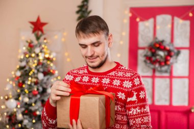 Noel ağacının önünde İskandinav desenli süveter giyen genç bir adam..