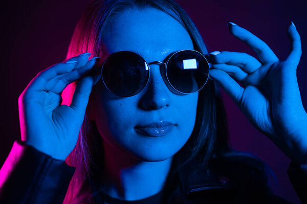 Cute girl with long hair near glowing pink neon city at night. Dyed blue pink hair of a young woman. Mysterious hipster teenager in round glasses. Cyberpunk light reflection.