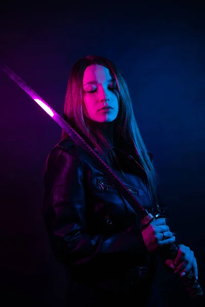 在黄昏时穿着黑色皮夹克的网络女孩拿着一个Katana 一个女人在俱乐部里拿着一把亚洲剑 手里拿着一支粉红色的蓝光 — 图库照片