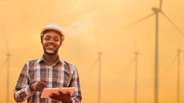 Afrikalı mühendis elinde tablet, rüzgar türbinlerinin önünde duruyor. Gün batımında elektrik santrali kuruyor..