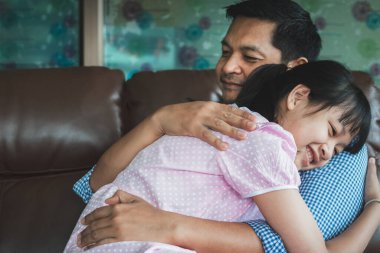 Baba ve kızı sevgiyle kucaklaşıyorlar, kameraya bakıyorlar ve evde kanepede otururken gülümsüyorlar. Babalar Günü kavramı.