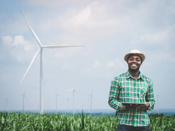 African farmer standing and holding digital tablet on corn farm with  wind turbine in background.Concept of green power sustainability resources  development by alternative energy
