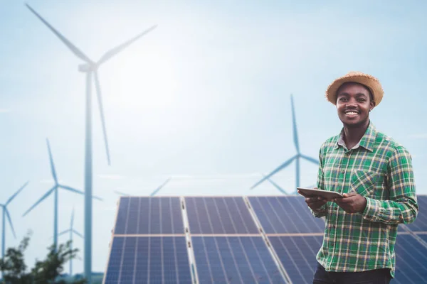 African farmer standing and holding digital tablet on corn farm with solar cell and wind turbine in background.Concept of green power sustainability resources  development by alternative energy