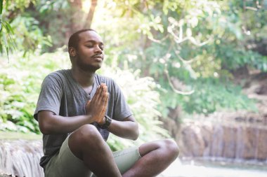 Tişört giyen Afrikalı bir adam şelalede oturup meditasyon yapar ve yoga egzersizleri yapar.