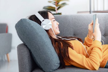 Cep telefonu ve kulaklık kullanan mutlu Asyalı kadın evdeki bir kanepede internet üzerinden müzik dinlerken medya eğlencesi rahatlıyor..