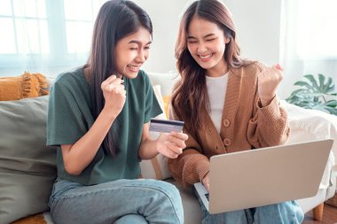 Two happy roommates shopping online together with credit card and laptop sitting on a sofa at home, Online payment.