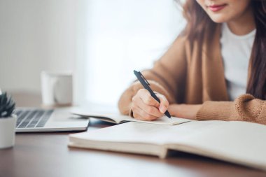 Close up - Hand of woman writing in spiral notepad placed on wooden desktop with various items at home. copy space.