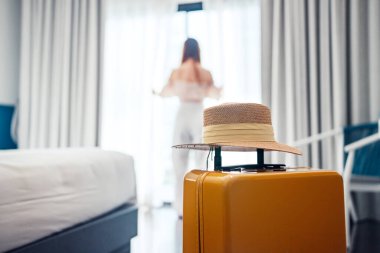 Close-up luggage with hat and blurred happy tourist woman background in hotel after check-in. Conceptual of travel and vacation.