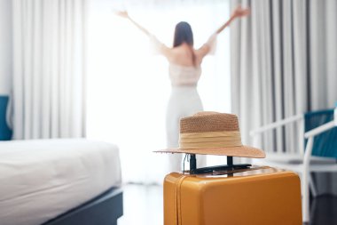 Close-up luggage with hat and blurred happy tourist woman background in hotel after check-in. Conceptual of travel and vacation.