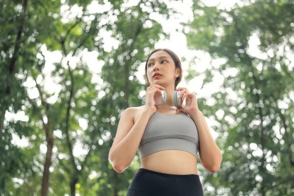 ジョギングを開始し 自然に囲まれたルート上の音楽を聴く前に無線ヘッドフォンを調整するアジアの女性を閉じます — ストック写真