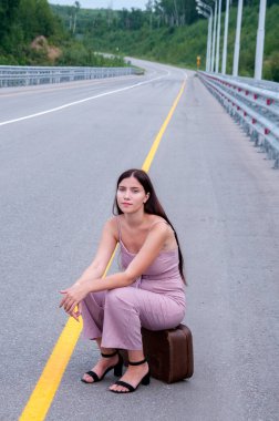 Bir kız boş bir yolun kenarında bir bavulda oturuyor ve araba bekliyor.
