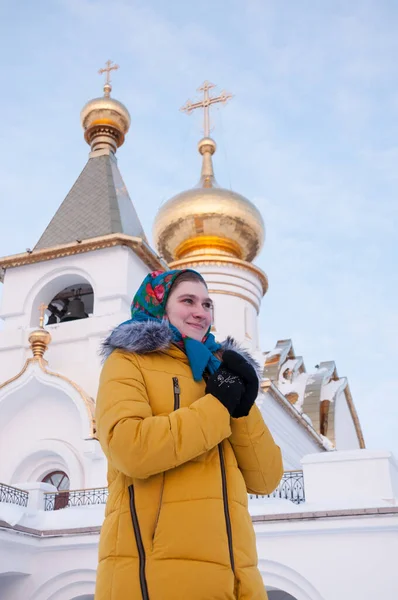 一个穿着俄罗斯式披肩和黄色羽绒服的女孩站在东正教教堂的背景下 — 图库照片