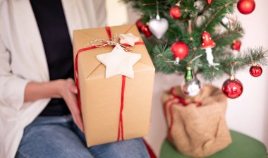 Female hands holding Christmas gift box with red bow and brown paper wrap for Xmas and New Year celebration party and decorating with the Christmas tree.
