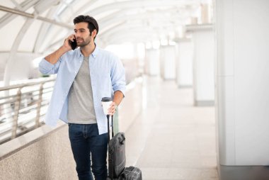 Adam akıllı telefonu kullanırken diğer elinde bir fincan kahve ve havaalanındaki bavulu çekiyor..