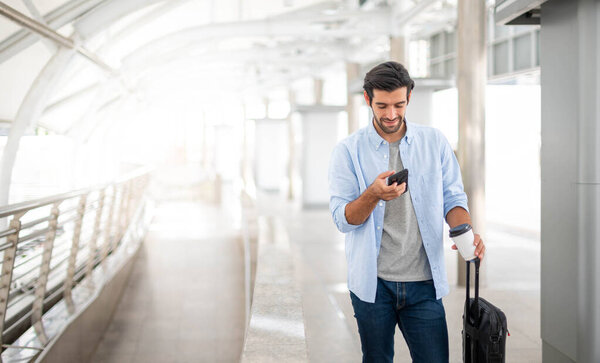 The man uses the smartphone while the other hand holding a cup of coffee and pulling the luggage at the airport.