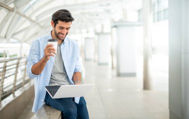 Kafkasyalı genç bir adam elinde bir fincan kahve tutuyor ve dış ofisteki taşınabilir bilgisayarı kullanarak serbest proje üzerinde çalışıyor..
