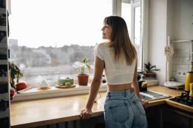 Çekici genç bir kadının portresi. Mutfakta dikilirken pencerenin yanındaki kameraya bakıyor.