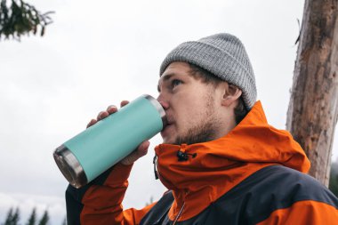 young man drinking water from thermos in winter forest