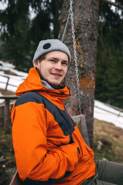 Turuncu ceketli ve ormanda gri şapkalı genç bir adam başka tarafa bakıyor. Gri şapkalı ve turuncu ceketli bir gencin portresi. Dağlarda bir salıncakta