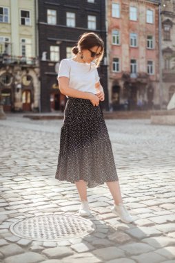 Yaz modası, beyaz tişört, benekli etek, beyaz spor ayakkabı ve güneş gözlüğü. Yazlık kıyafetli şık bir kız eski kasaba sokaklarında yürüyor.