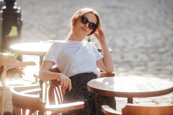 Güneş gözlüklü, beyaz tişörtlü ve puantiyeli etekli şık bir kız bir kafede dinleniyor. Genç kadın portresi