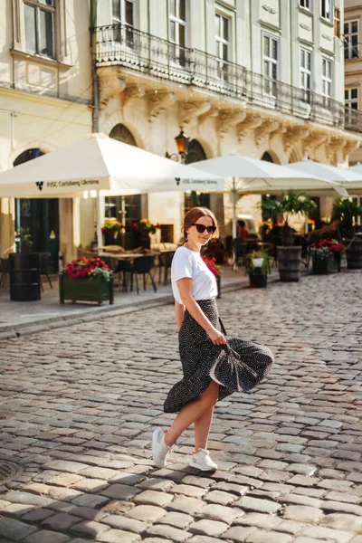 Yazlık giysiler içinde şık bir kız. Eski şehrin arka planında beyaz tişörtlü ve benekli etekli kadın portresi