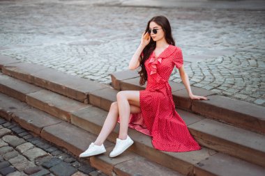 Yaz modası: kırmızı benekli elbise ve beyaz spor ayakkabılar. Merdivenlerde retro kıyafetli güzel kız. Yaz mevsimi modası