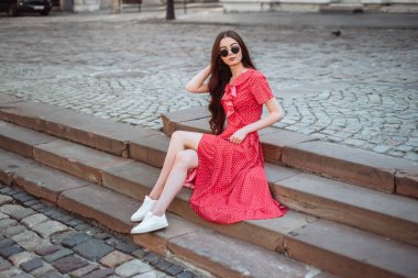 Yaz kıyafetleri: kırmızı benekli elbise ve beyaz spor ayakkabılar. Esmer kız eski kasabanın merdivenlerinde oturuyor. Yaz portresi