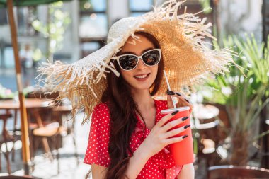 Sıcak ve güneşli bir günde soğuk içecek. Güneş gözlüklü, saman şapkalı, rahat genç bir kadının yaz portresi.