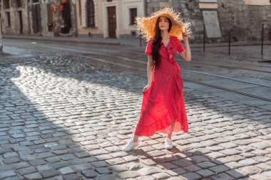 Yazlık kıyafet: hasır şapka, güneş gözlüğü, kırmızı benekli elbise ve beyaz spor ayakkabılar. Şık kız eski şehir sokaklarında yürüyor