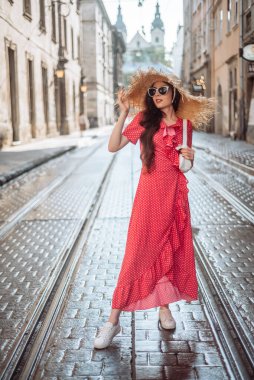 Yazlık kıyafet: hasır şapka, güneş gözlüğü, kırmızı elbise, beyaz çanta ve beyaz spor ayakkabılar. Şık kız eski şehir sokaklarında yürüyor