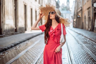Yazlık kıyafetli şık kız: hasır şapka, güneş gözlüğü, kırmızı elbise, eski şehir sokaklarında beyaz çanta yürüyüşü