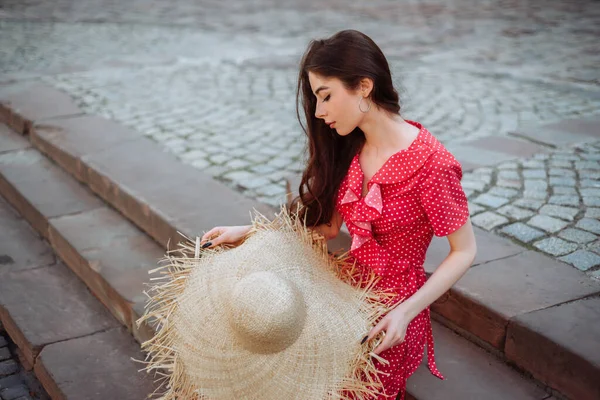 身穿红圆点裙 头戴草帽的时髦姑娘坐在镇广场的楼梯上 年轻女子户外夏季肖像 — 图库照片