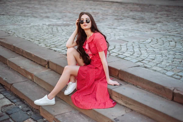 穿着红色圆点裙和白色运动鞋的漂亮女孩坐在镇广场的楼梯上 年轻女子户外夏季肖像 — 图库照片