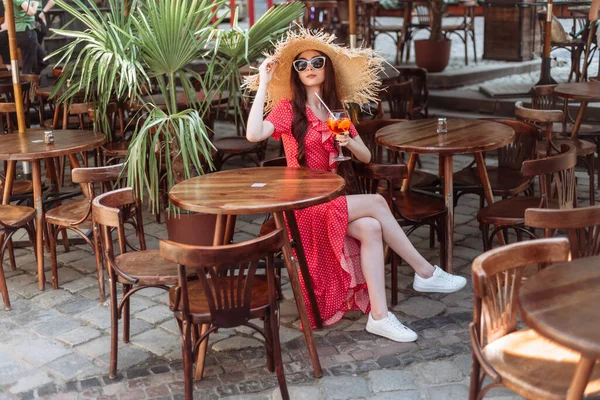 Tarz sahibi bir kız terasta soğuk yaz kokteyli aperol spreyi içer. Güneş gözlüklü, hasır şapkalı ve kırmızı benekli genç bir kadının yaz dönemi portresi