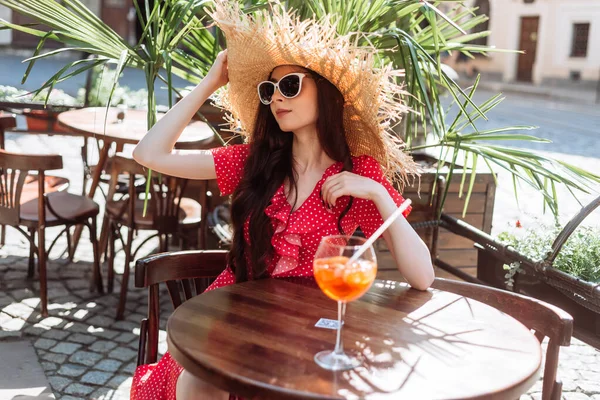 Tarz sahibi bir kız terasta soğuk yaz kokteyli aperol spreyi içer. Güneş gözlüklü, hasır şapkalı ve kırmızı benekli genç bir kadının yaz dönemi portresi