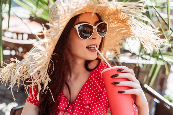 Güneşli terasta soğuk yaz içkisi içen şık bir kız. Güneş gözlüklü, hasır şapkalı ve kırmızı benekli genç bir kadının yaz dönemi portresi