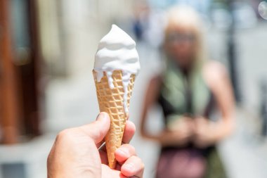 Ön planda, bir adamın eli beyaz dondurmalı waffle külahını tutuyor, eriyip içine damlıyor..