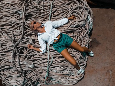 Beyaz gömlekli, mavi şortlu ve güneş gözlüklü genç bir kız bir yığın ipin üzerinde yatıyor. İnsansız hava aracı fotoğrafı..