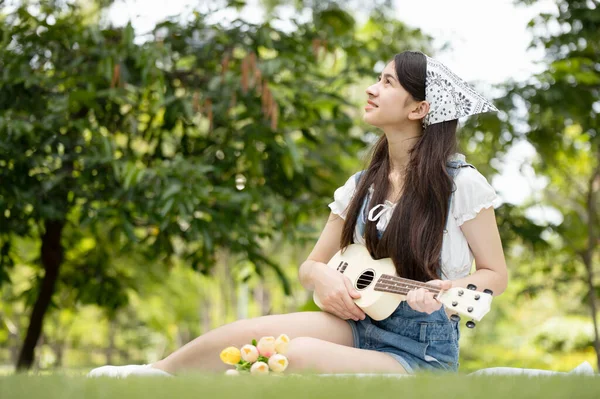 Genç Asyalı kadın parkta Ukulele çalarak eğleniyor ve dinleniyor. Müzik zamanı.