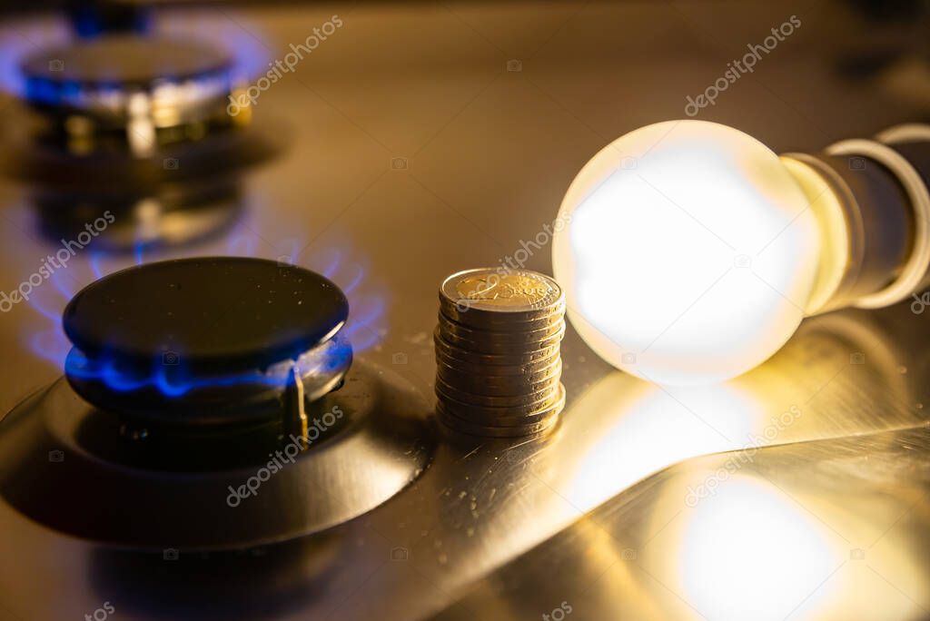 Light bulb on next to lit gas cooker, with coins next to it. Energy and gas costs, cost increases.Light bulb on next to lit gas cooker, with coins next to it. Energy and gas costs, cost increases.