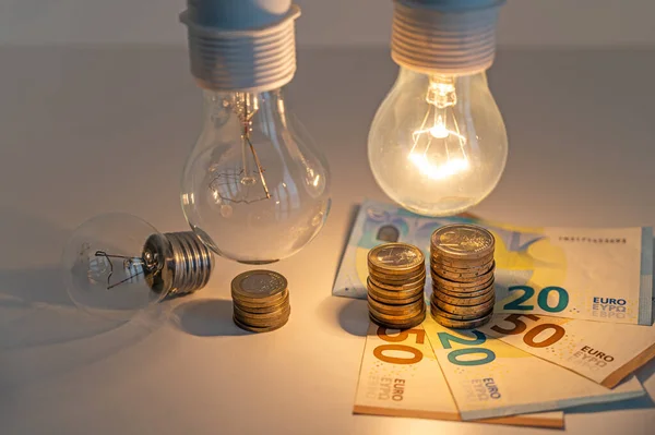 Light bulbs, one on and one off, above a pile of coins of increasing height. Increases in energy tariffs.