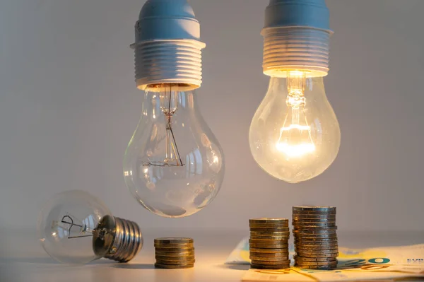 Light bulbs, one on and one off, above a pile of coins of increasing height. Increases in energy tariffs.