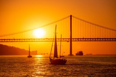 Sailboat with the Ponte 25 de Abril or 25the April Bridge at the Rio Tejo near the City of Lisbon in Portugal.  Portugal, Lisbon, October, 2021