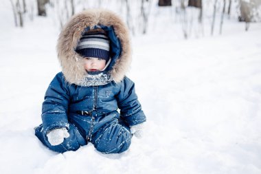 Ladin ormanında karda oturan bir çocuğun portresi. Küçük çocuk kışın dışarıda eğleniyor. Noel tatili. Mavi tulumlu, örgü örüp parkta yürüyen şirin bir çocuk..
