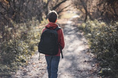 Sonbahar parkında sırt çantalı genç bir çocuk yolda yürüyor. Aktif yaşam tarzı, okula dönüş. Sonbahar ormanındaki öğrenci..