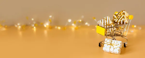İnternetten noel alışverişi ve indirimi. Altın arka planda ışıklı hediye kutularıyla alışveriş arabası. Süpermarket tramvayı tam hediyeler. Kopyalama alanı olan pankart