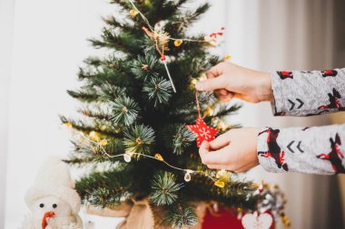 Noel ve yeni yıl tatili. Küçük Noel ağacını süslemek için çocuk elleri yakın plan. Pijamalı çocuk köknar dallarına ahşap oyuncaklar asıyor. Kış tatili yaşam tarzı.